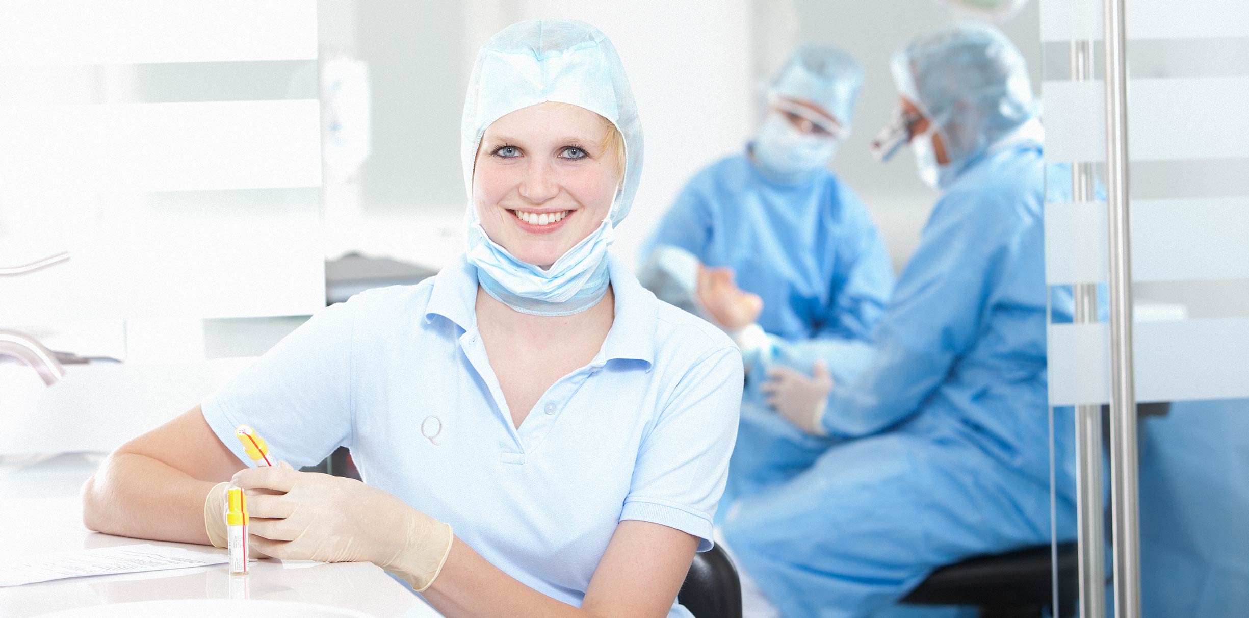 Eine junge Zahnarzthelferin lächelt im Zahn-Op-Saal in die Kamera. Die Zahnärzte reden im Hintergrund mit der Patientin.